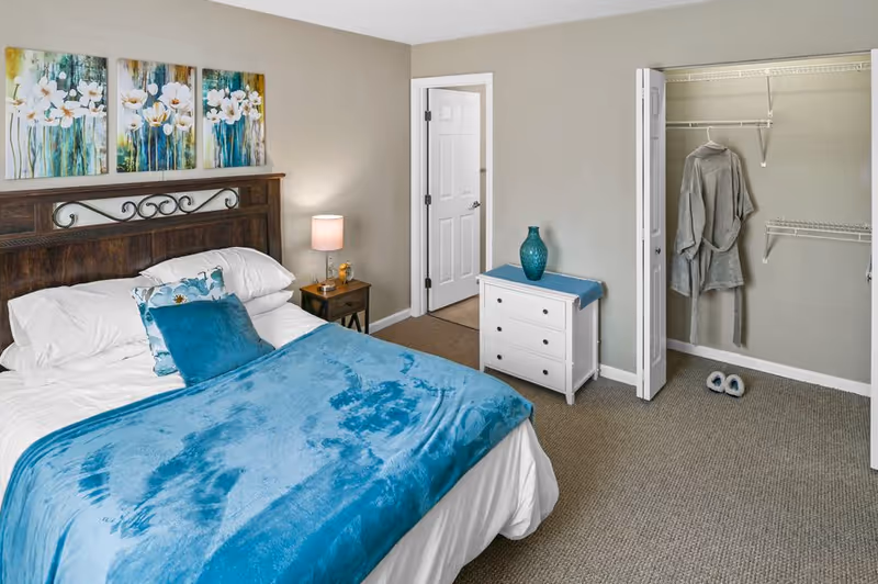 A neatly arranged bedroom with a wooden headboard bed covered with white sheets and a blue blanket. There are two pillows on the bed, one white and one blue. A nightstand with a lamp and a small decorative item is beside the bed. On the wall above the bed, there are three floral paintings. Across from the bed, there is a white dresser with a blue vase on top and an open closet with a gray robe hanging inside and a pair of slippers on the floor. The walls are painted light gray and the floor is carpeted.