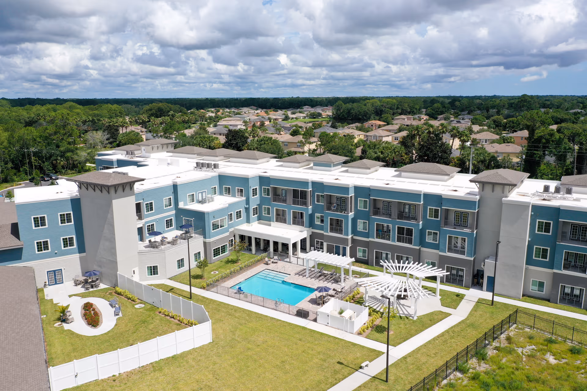 Aerial view of a three-story senior living facility building with blue and gray exterior walls, a central outdoor swimming pool surrounded by lounge chairs, white pergolas, and landscaped green areas. The building is situated in a suburban neighborhood with many houses and trees in the background under a partly cloudy sky.