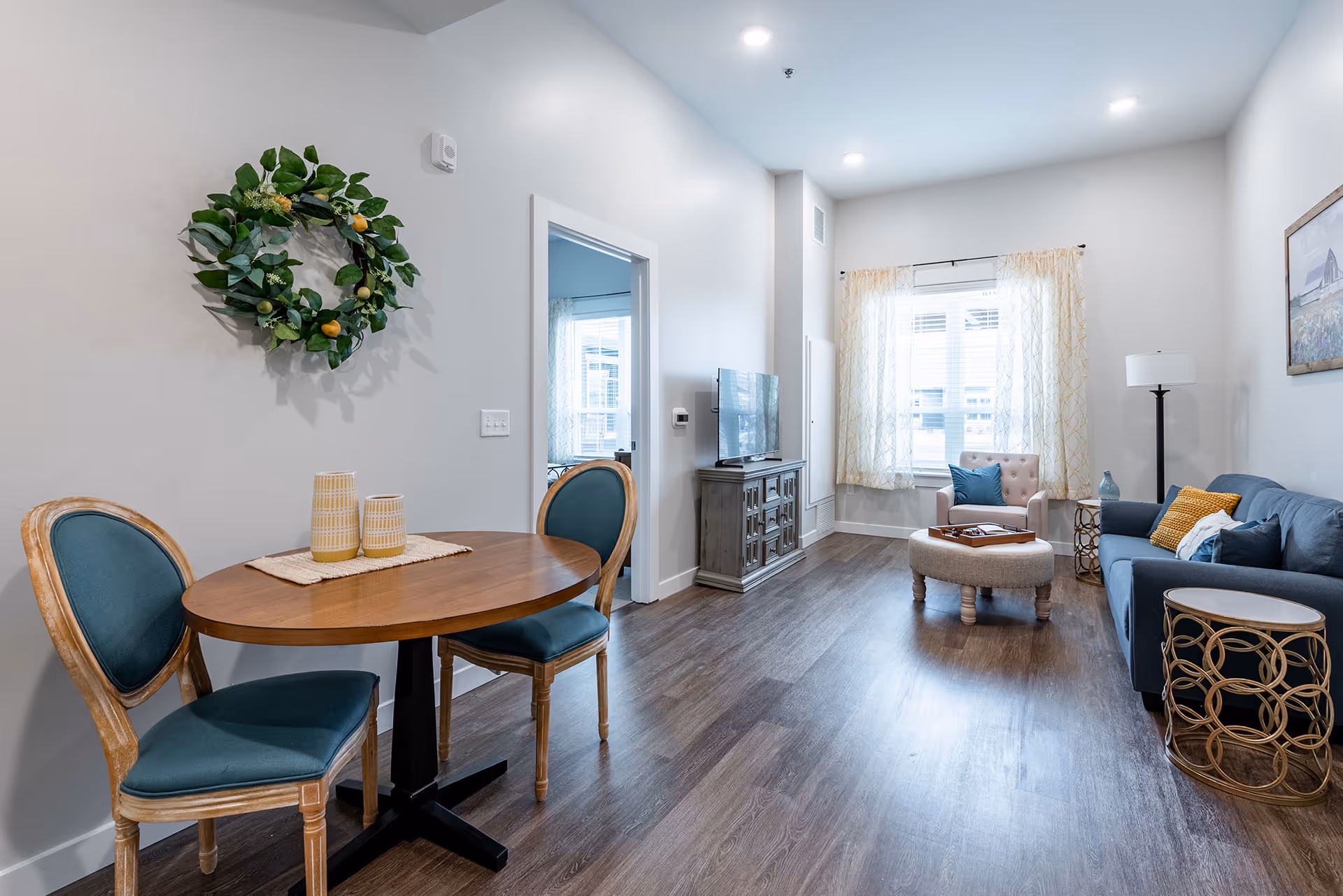 Bright modern living room and dining area with a round wooden table and teal chairs, sofa, armchair, TV stand, and a window with sheer curtains.