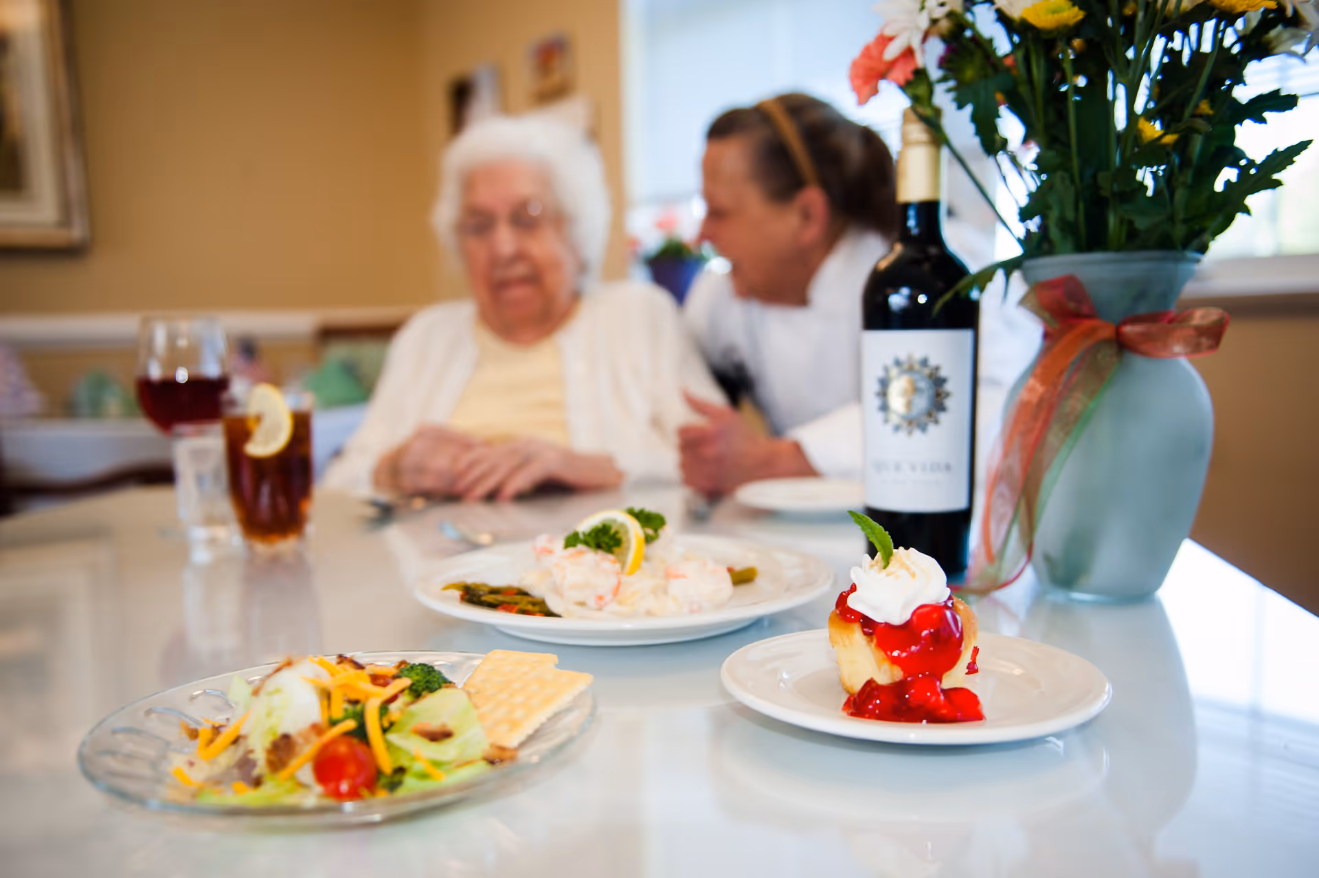 A dining table with plates of food including a salad with crackers, a shrimp dish, and a dessert topped with whipped cream and cherry sauce. A bottle of wine and a vase with flowers are also on the table. In the background, an elderly woman and a caregiver are engaged in conversation.