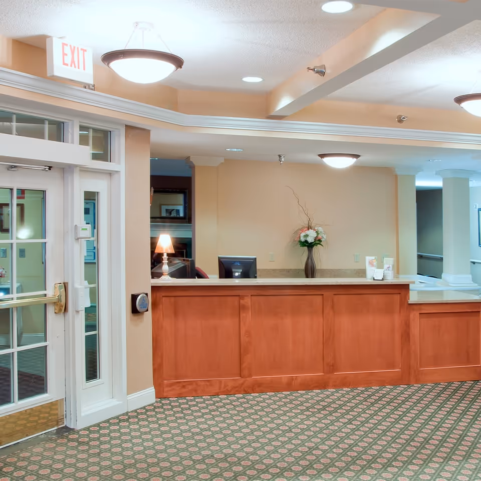 Reception area with a wooden front desk, a computer monitor, a lamp, and a vase with flowers. To the left, there is a glass door with an exit sign above it. The floor is carpeted with a patterned design, and the walls are painted in a warm beige tone.