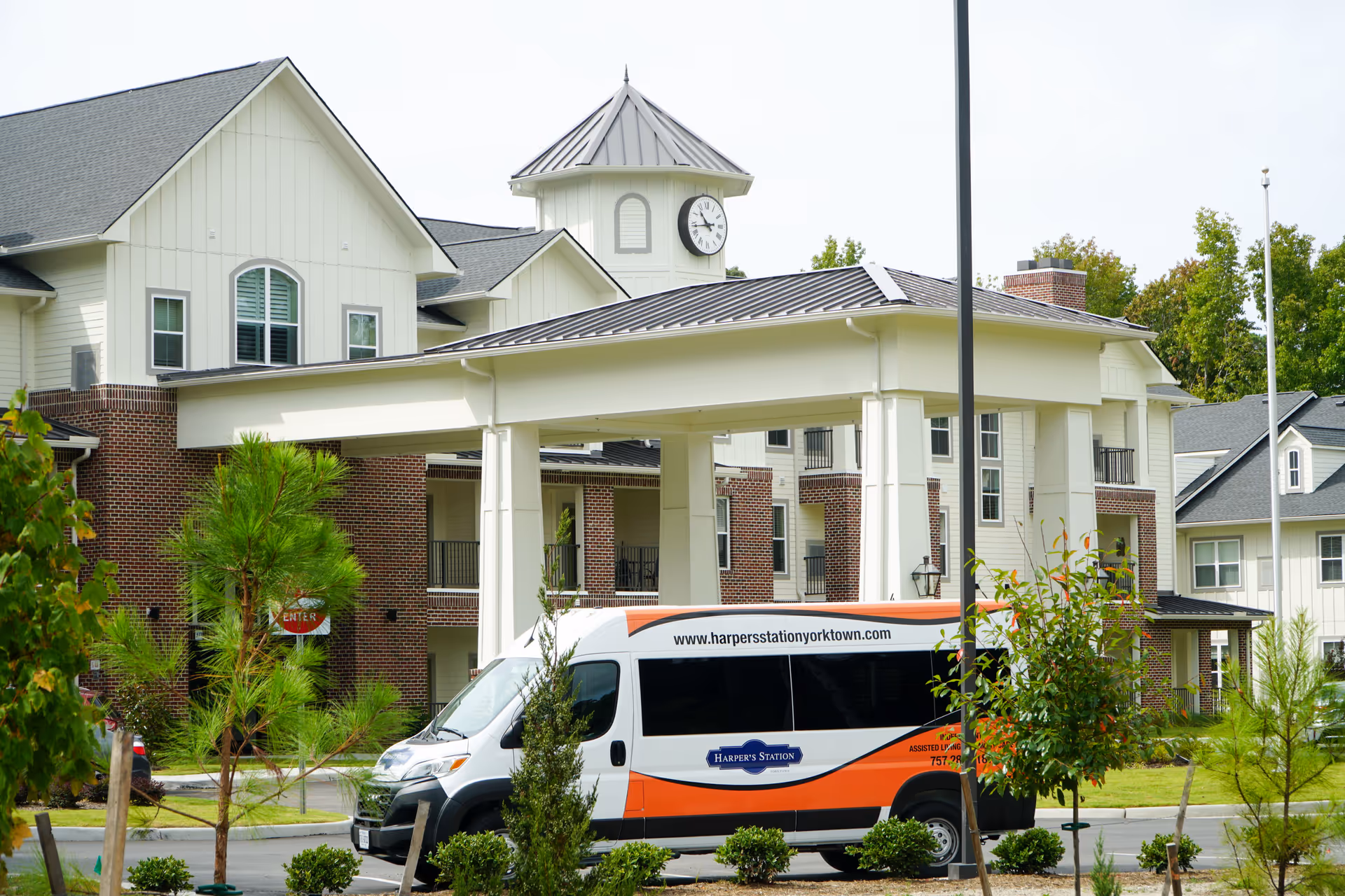 Exterior view of Harper's Station Yorktown senior living facility showing a covered entrance with a clock tower above it. A white and orange shuttle van with the facility's name and website is parked in front. The building features a combination of brick and light-colored siding with multiple windows and balconies. Trees and shrubs are planted around the driveway and entrance area.