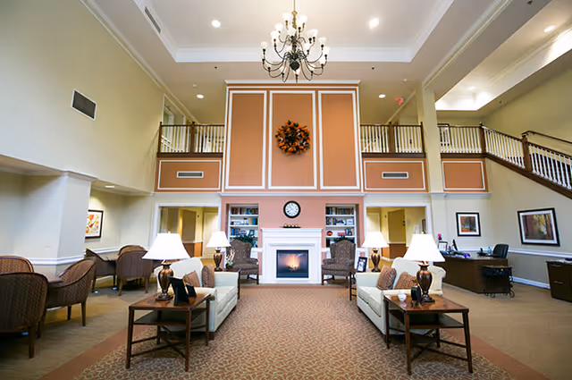 Spacious and well-lit living room area with high ceilings, a chandelier, and a fireplace centered on a wall with decorative paneling and a wreath. The room features two sofas facing each other with side tables and lamps, armchairs, framed artwork on the walls, and a desk area on the right side.