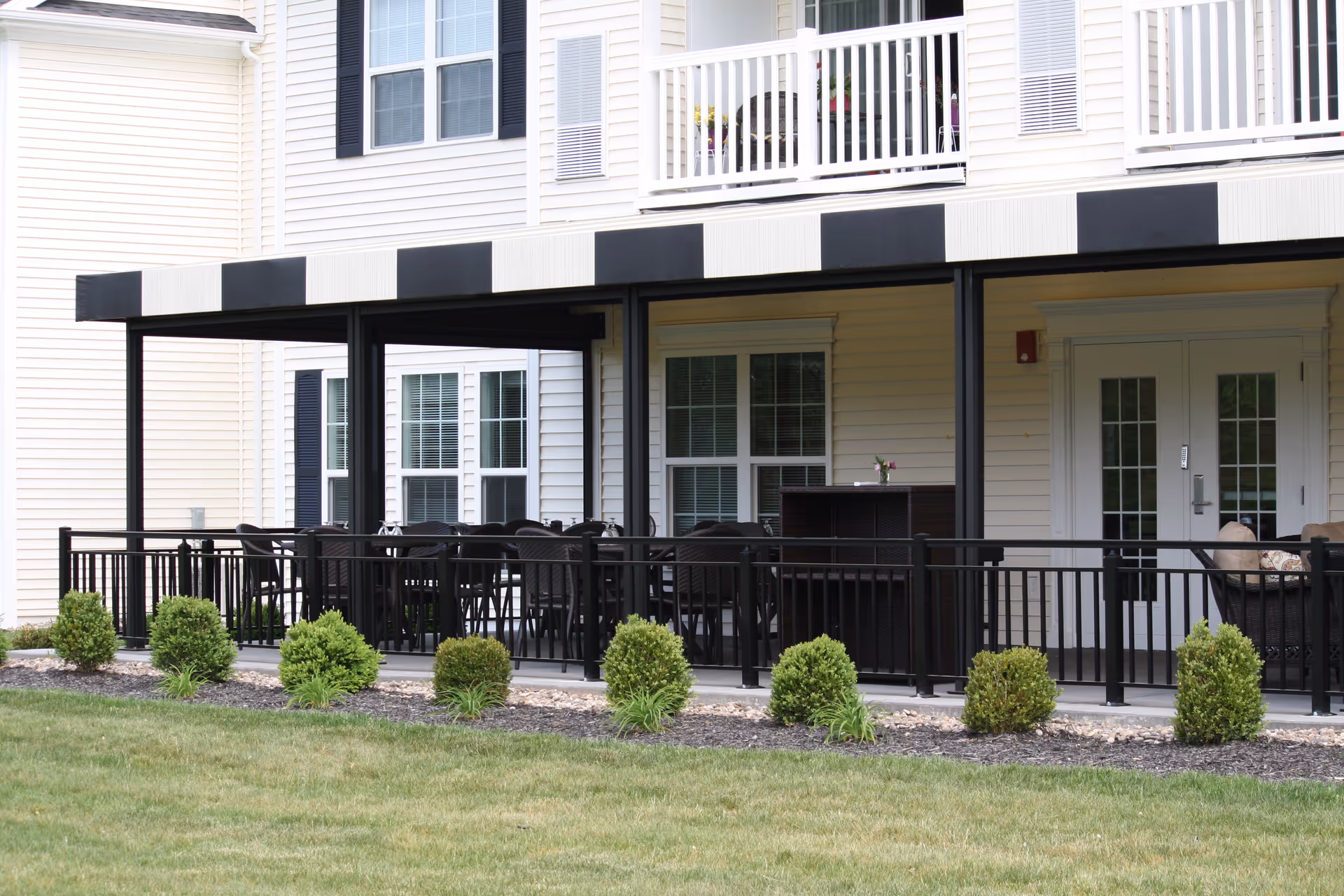 Outdoor covered patio area at a senior living community with black metal railing, black and white striped awning, multiple black chairs and tables, and small green bushes planted in front of the patio. The building exterior is light-colored with white-framed windows and doors.