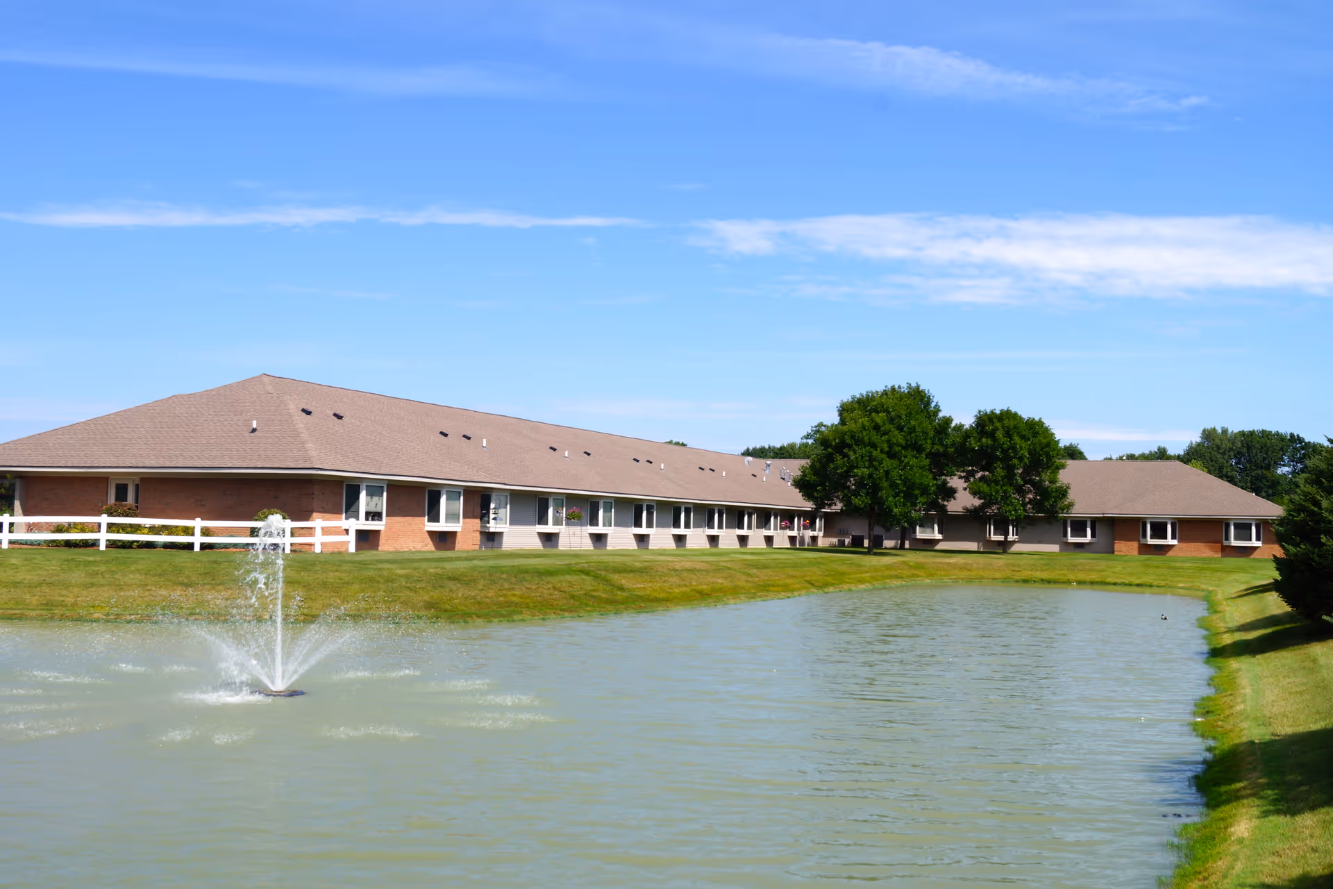 A single-story brick building with multiple windows, surrounded by green grass and trees, next to a pond with a water fountain under a clear blue sky.