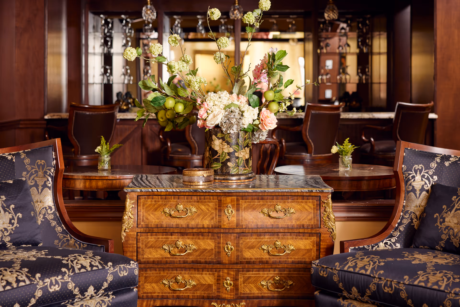 A richly decorated lounge with a wooden chest topped by a floral arrangement between two patterned armchairs and a mirrored bar in the background.