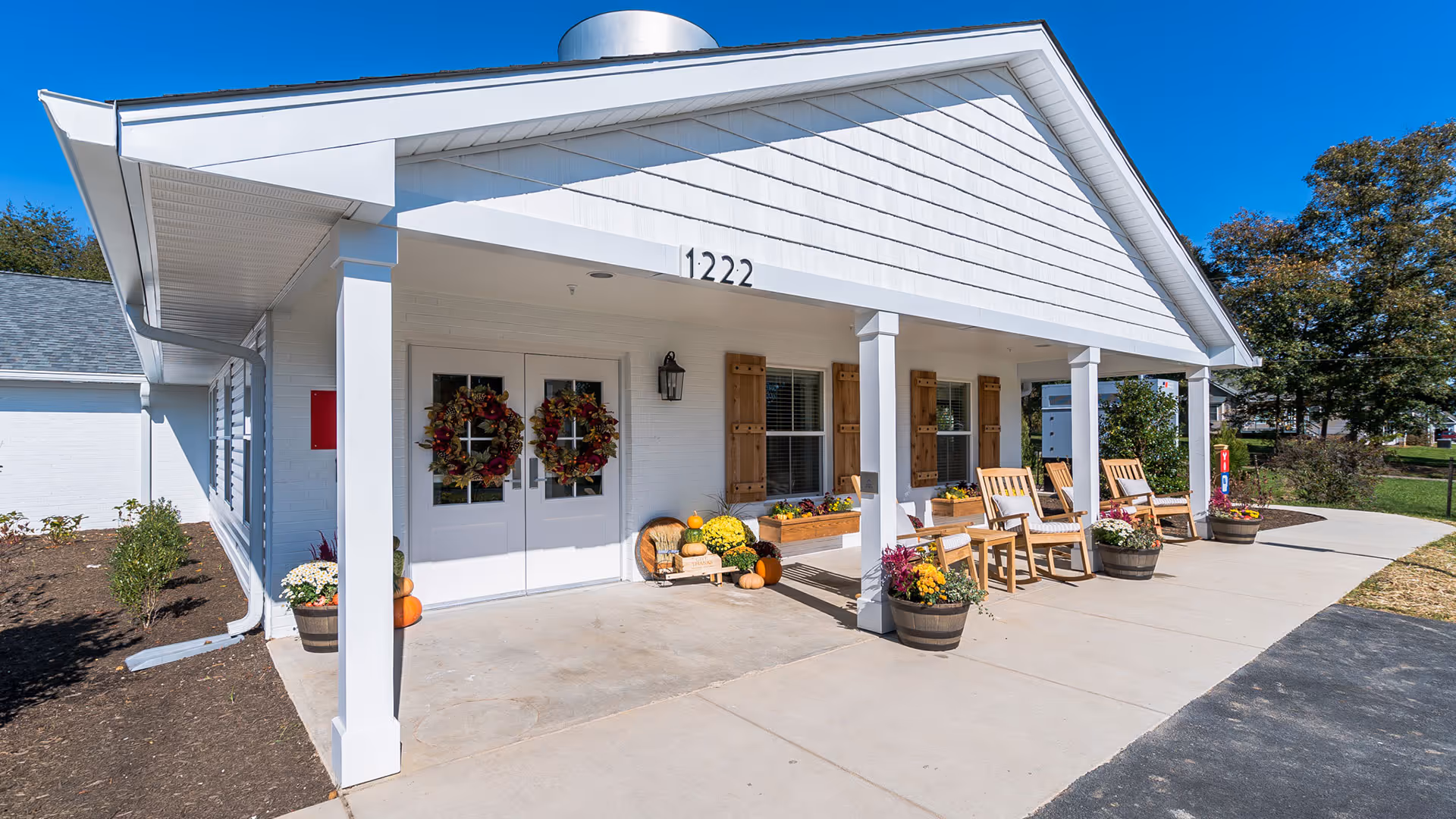 Front entrance of a white building with a covered porch, double doors decorated with wreaths, wooden chairs, and potted plants.