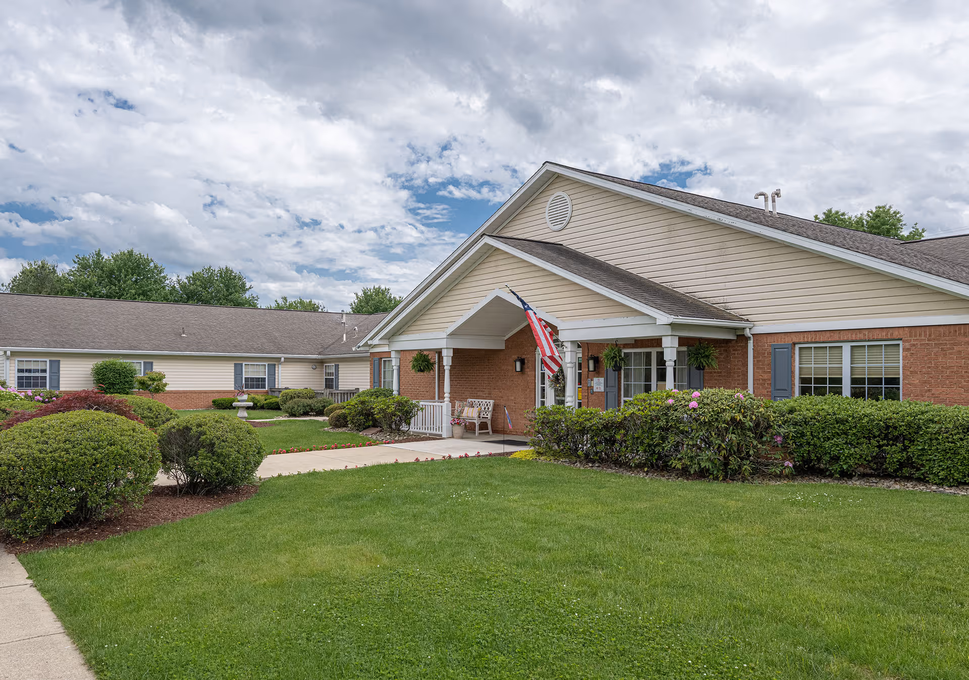 Exterior view of Arden Courts - ProMedica Memory Care Community in Monroeville, showing a single-story building with beige siding and brick accents, a covered entrance with an American flag, well-maintained green lawn, bushes, and a partly cloudy sky.