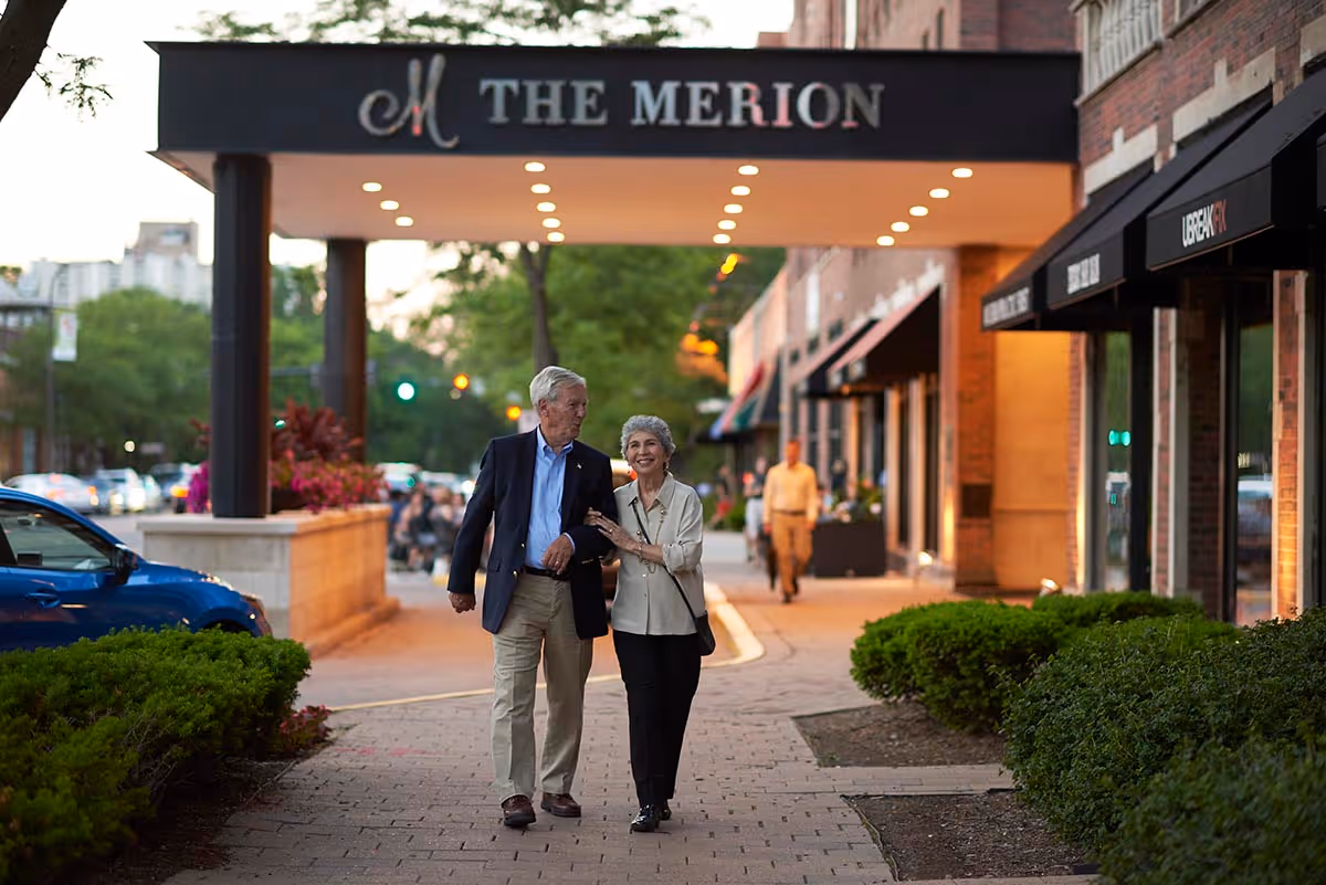 An elderly couple stroll arm-in-arm along a sidewalk beneath a marquee reading "The Merion" at the building entrance.