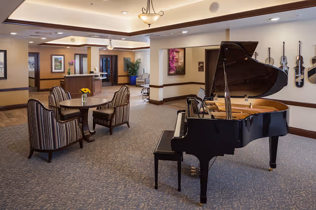 A spacious senior living common area featuring a black grand piano with its lid open on the right side. To the left, there is a round wooden table with four striped upholstered chairs around it and a small vase with yellow flowers on the table. The room has beige walls with dark wood trim, carpeted flooring, framed artwork on the walls, and ceiling lights providing warm illumination. In the background, there is a reception desk and additional seating areas with plants and decorative elements.