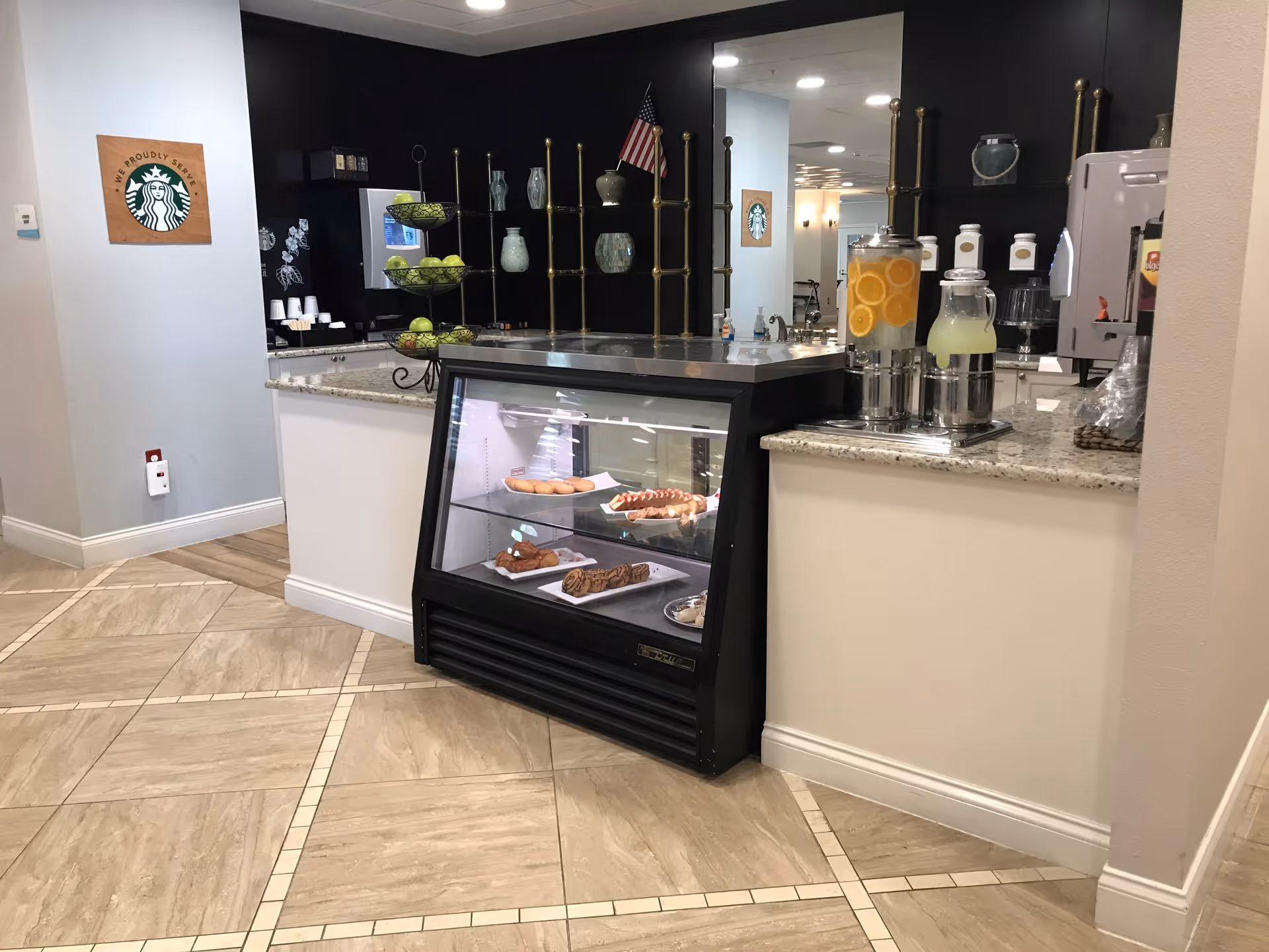 A small cafe area inside a senior living facility with a glass display case containing pastries, a countertop with beverage dispensers filled with lemon water and lemonade, a tiered fruit stand with green apples, and a Starbucks logo sign on the wall.