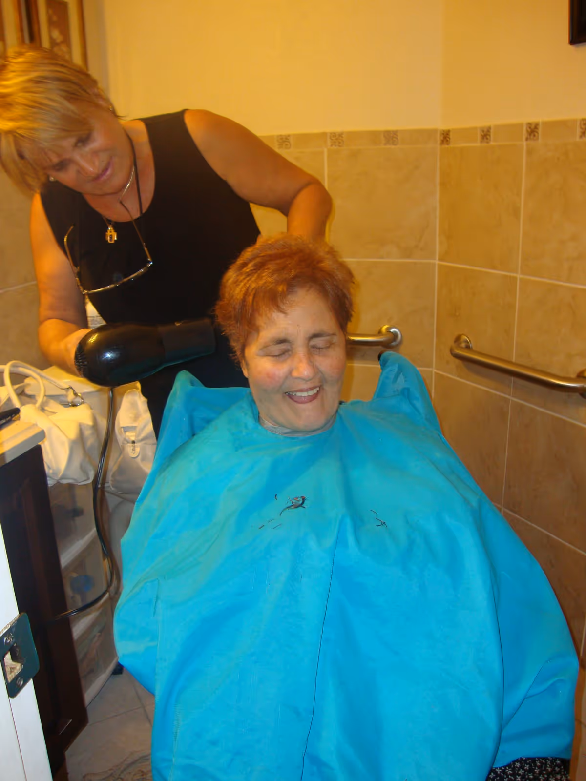 A woman is drying the hair of an elderly woman who is seated and covered with a blue cape in a tiled room with grab bars on the walls.