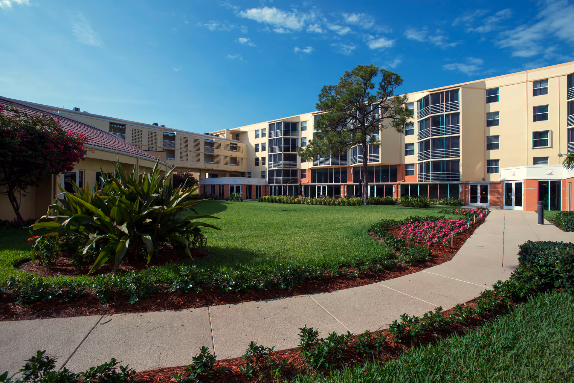 Courtyard with lawn, flowerbeds and a walkway in front of a multi-story senior living building under a blue sky.