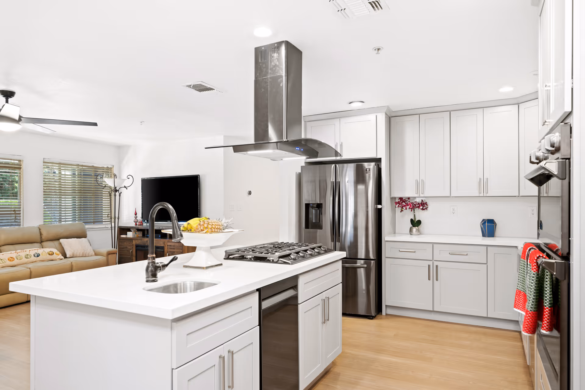 Bright and modern kitchen with white cabinetry, a large island with a sink and gas stove, stainless steel refrigerator, and oven. In the background, a living area with a beige sofa, a TV on a wooden stand, and windows with blinds is visible.
