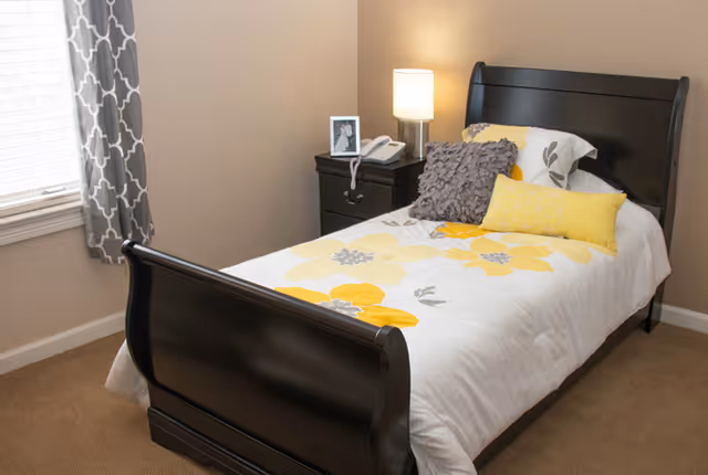 A bedroom with a single dark wooden sleigh bed featuring white bedding with large yellow and gray floral patterns. There are three decorative pillows on the bed in gray and yellow. Next to the bed is a dark wooden nightstand with a lamp, a telephone, and a framed photo. A window with gray and white patterned curtains is visible on the left side.