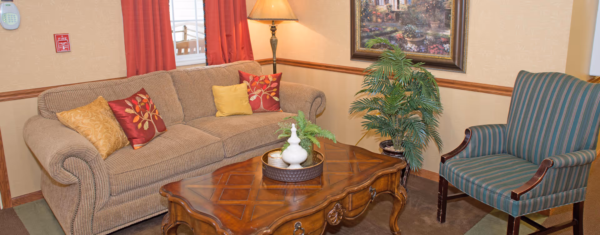 A cozy living room area with a beige sofa adorned with red and yellow decorative pillows, a wooden coffee table with a small plant and decorative items, a striped armchair, a floor lamp, a potted plant, and a framed painting on the wall.