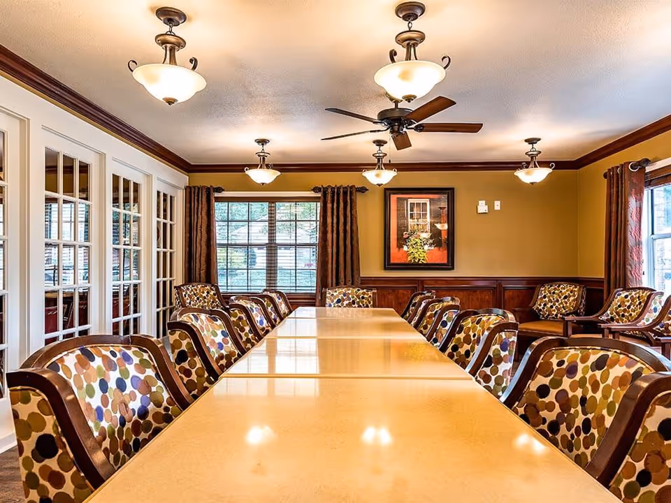Long dining table surrounded by patterned chairs in a well-lit senior living dining room with pendant lights, ceiling fan, and windows.