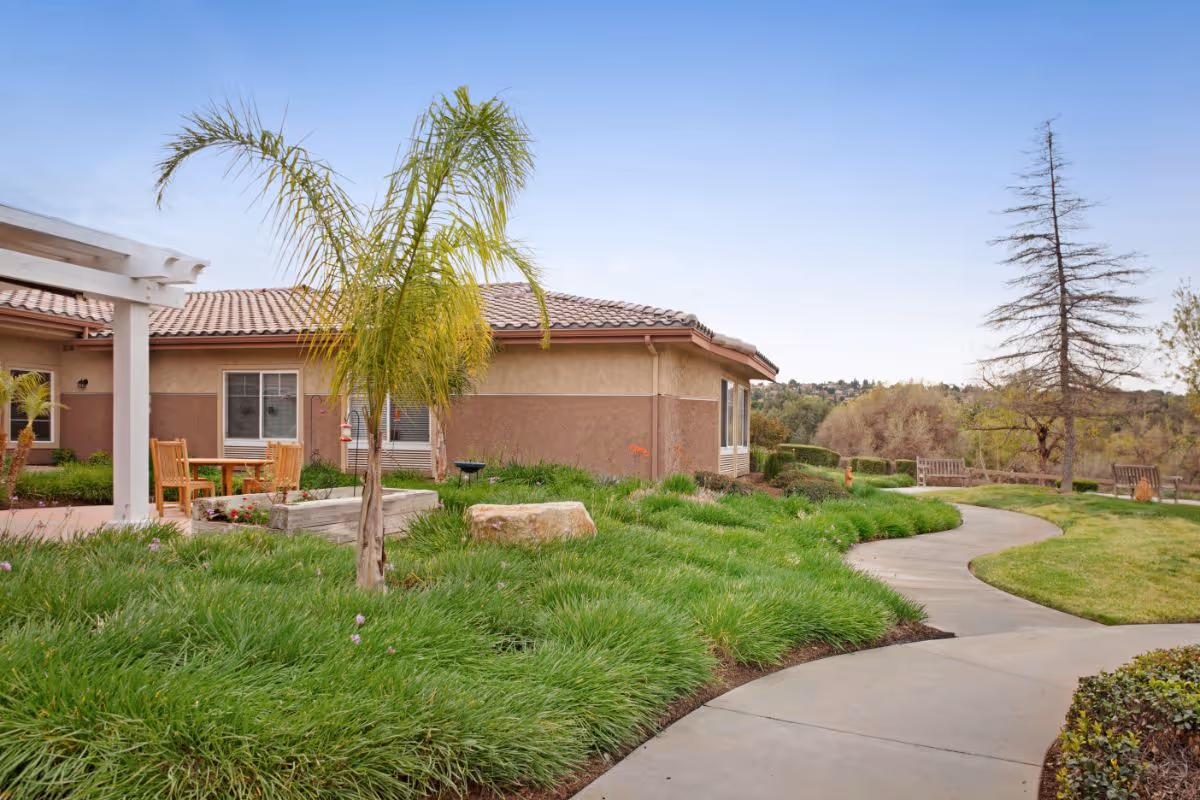 Curved walkway through a landscaped courtyard with a palm tree, seating, and a single-story stucco building under a blue sky.