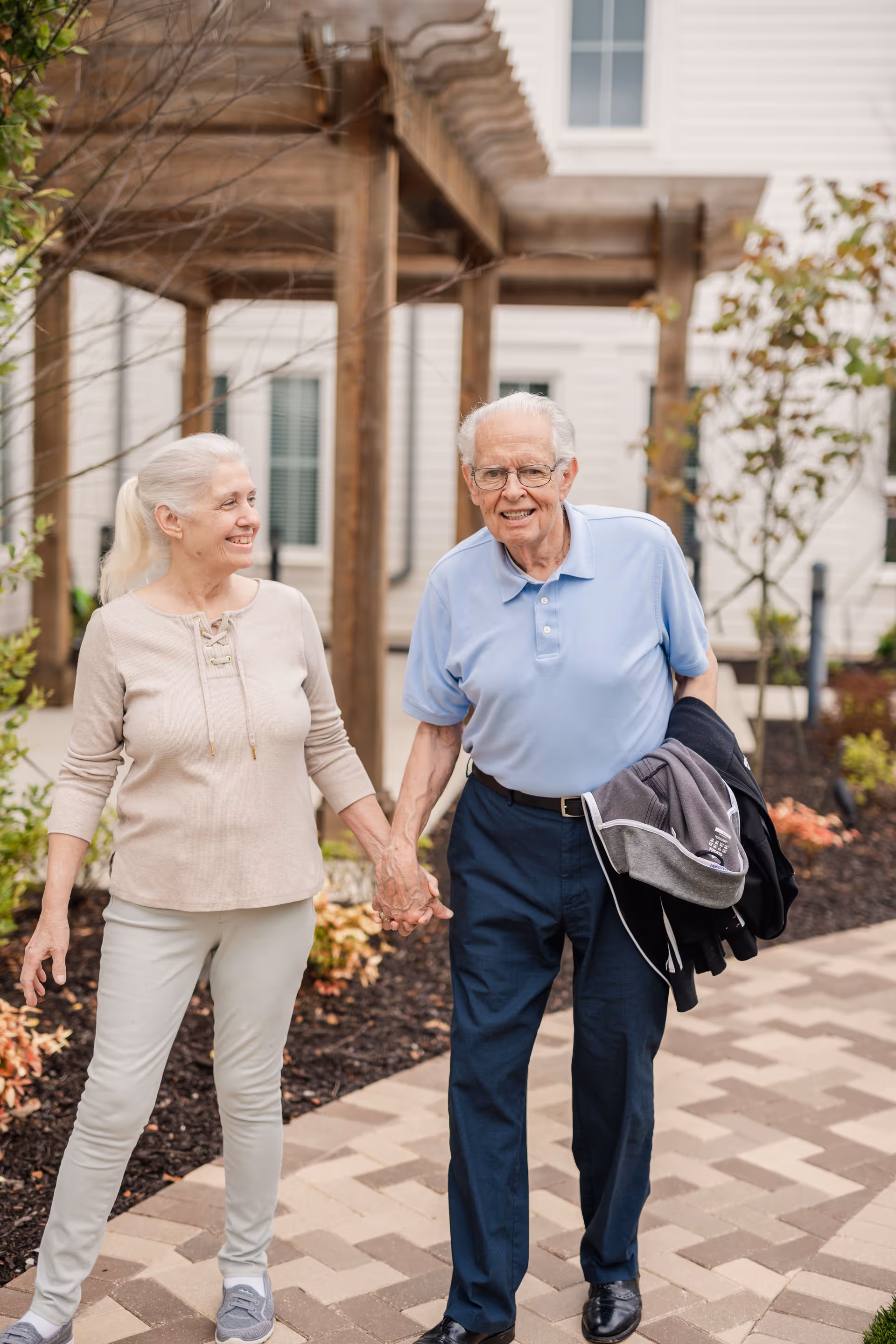 An elderly couple holding hands and walking outside on a paved pathway near a wooden pergola and landscaped garden beds. The woman is wearing a beige long-sleeve top and light pants, while the man is wearing a light blue polo shirt and dark pants, carrying jackets in his arm. They are smiling and appear happy.