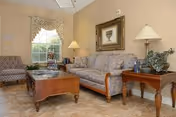 A cozy living room with a floral patterned sofa, a wooden coffee table, and an armchair near a window with a valance. There are two table lamps on wooden side tables, a framed picture on the wall, and a small plant on one of the tables. The room has beige walls and tiled flooring.