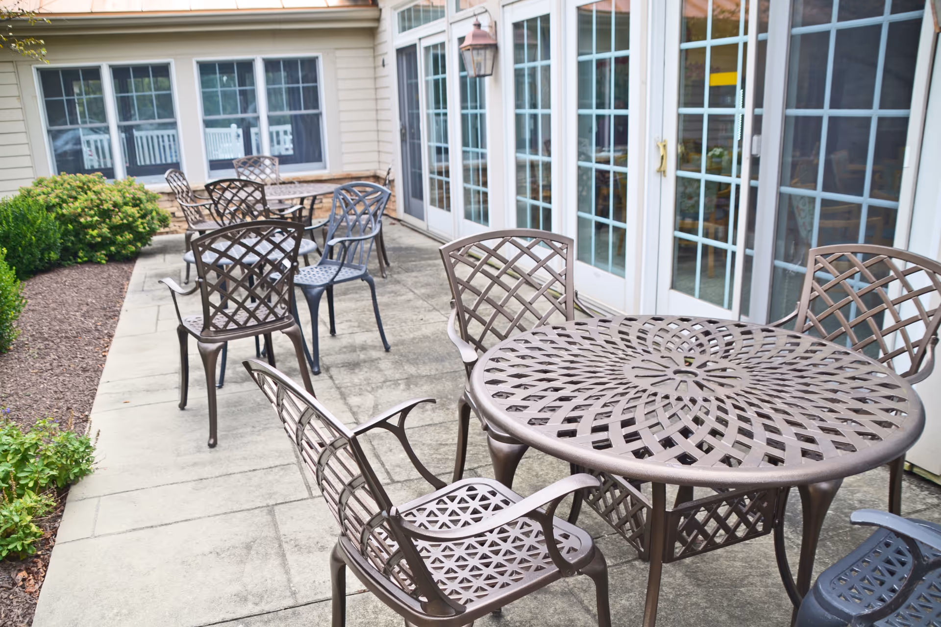 Outdoor patio area with multiple metal tables and chairs arranged on a concrete surface next to a building with large glass doors and windows. There are bushes and plants along the edge of the patio.