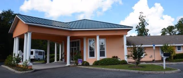 Exterior view of a single-story building with a covered entrance supported by white columns, surrounded by greenery and a clear blue sky with clouds.