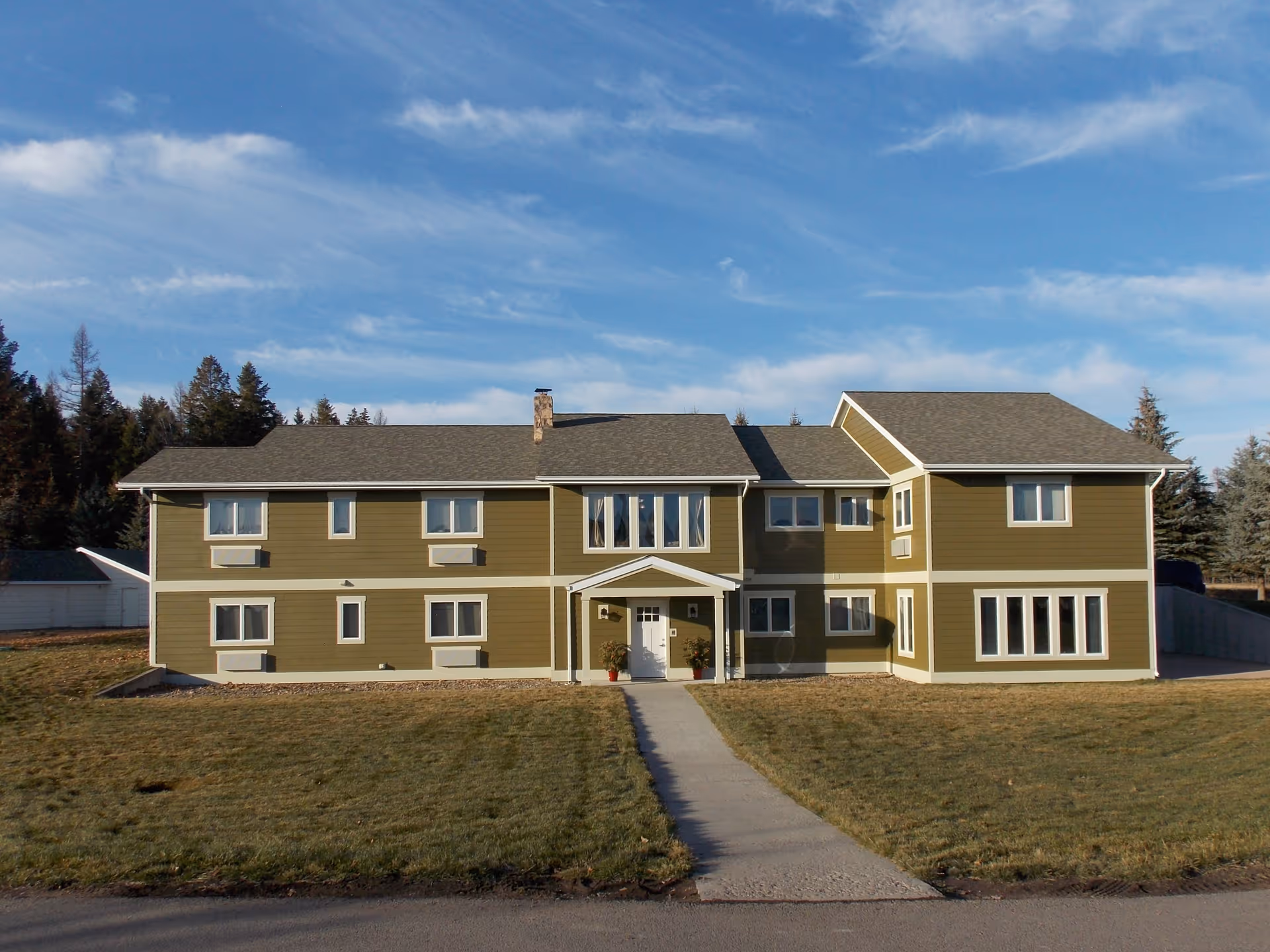 Front exterior of a two-story olive-green residential building with a central entrance, walkway, and lawn under a blue sky.