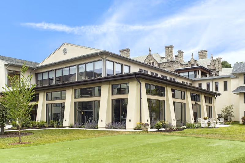 Exterior view of a senior living facility building with large windows, beige walls, and a well-maintained green lawn in front. The sky is clear with some clouds, and there are some small trees and shrubs around the building.