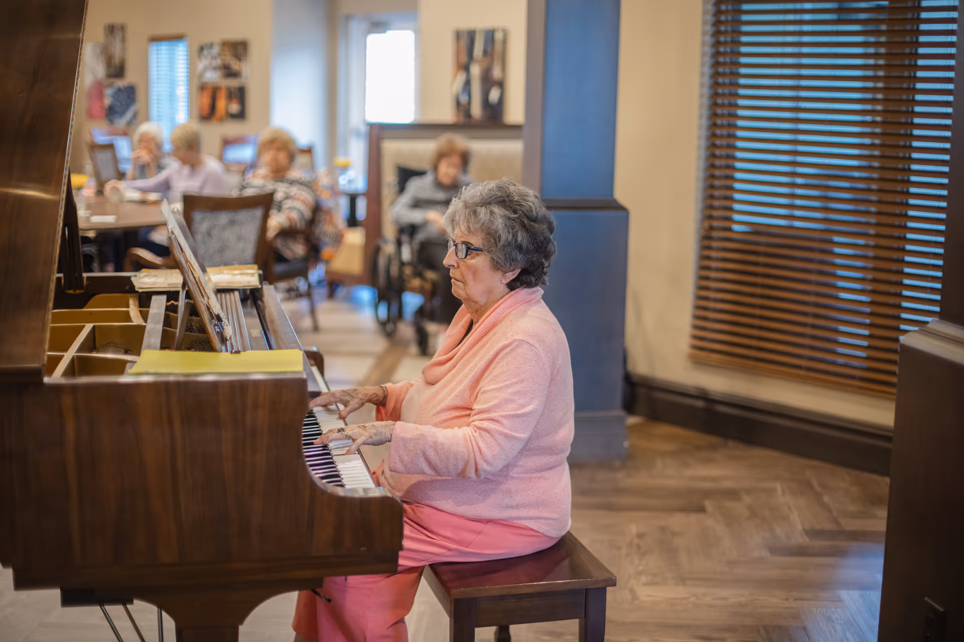 An elderly woman wearing glasses and a pink sweater is playing a wooden piano in a senior living facility. In the background, several other elderly individuals are seated at tables, some in wheelchairs, in a well-lit room with wooden blinds on the windows.