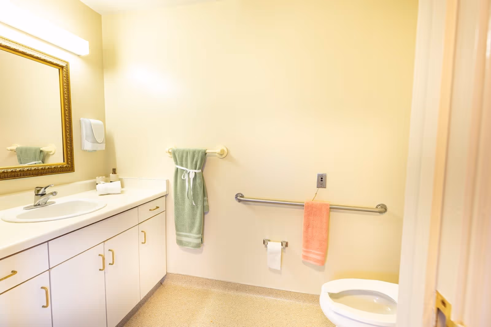 A clean senior living facility bathroom with a white sink and countertop, a large mirror with a gold frame, a green towel hanging on a wall-mounted towel rack, a pink towel hanging on a grab bar, and a white toilet partially visible on the right side.