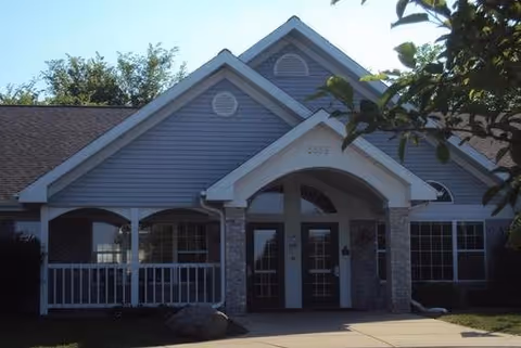 Front exterior view of a single-story building with a peaked roof, blue siding, and brick accents. The entrance features double glass doors under an arched portico supported by brick columns. There is a small porch area with white railing to the left of the entrance, and trees partially frame the building.