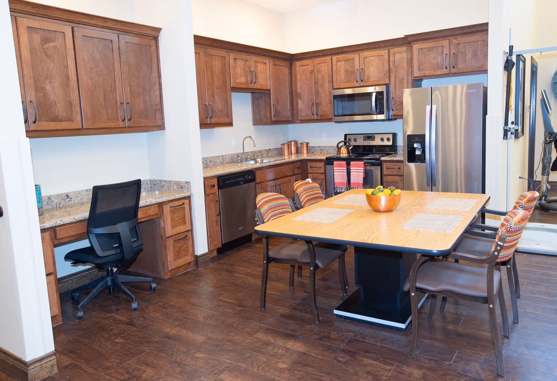 Bright communal kitchen and dining area with wooden cabinets, stainless steel appliances, a central wooden table with chairs, and a small desk workspace.