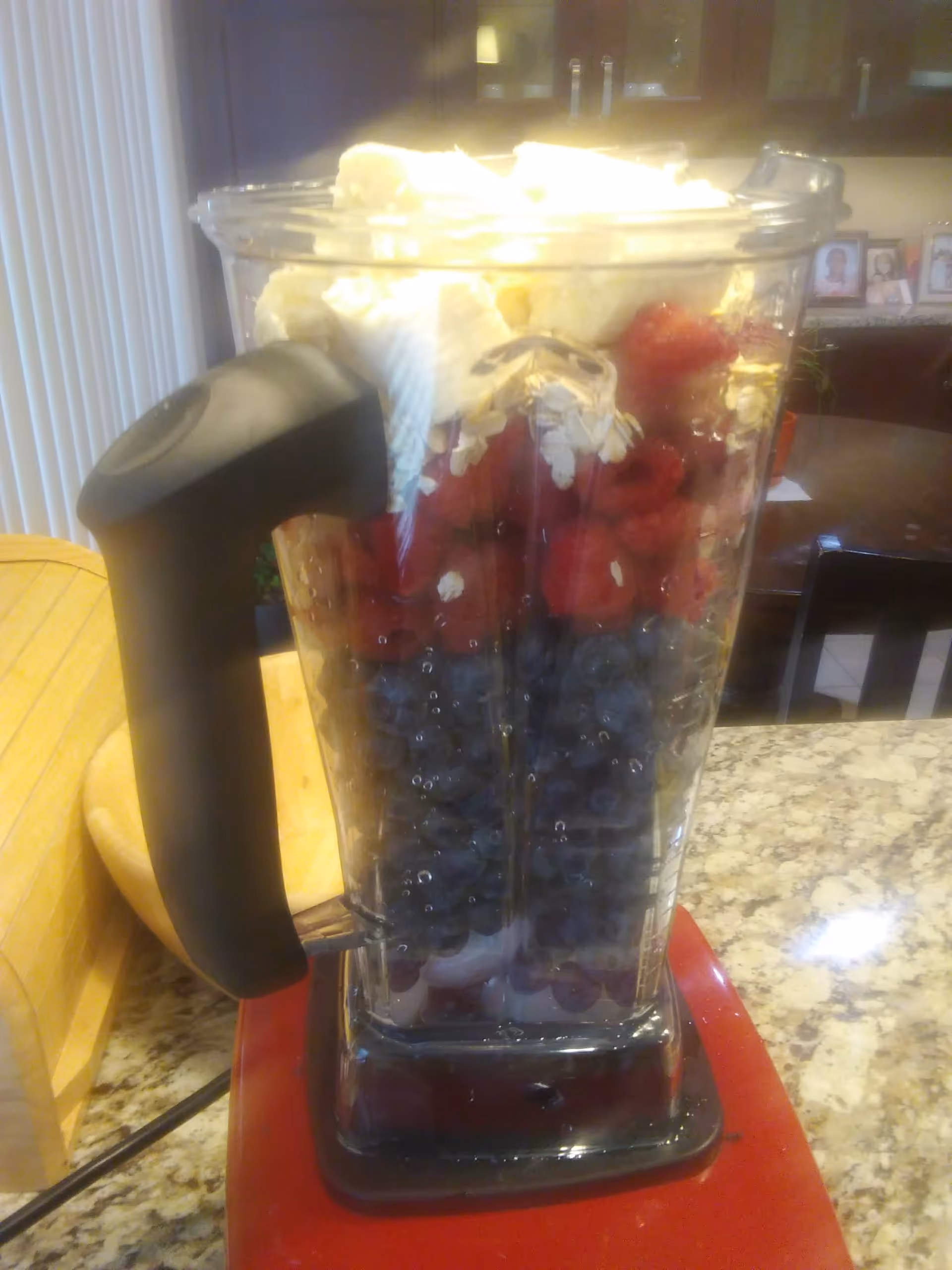 A blender filled with layers of blueberries, raspberries, oats, and banana slices on a kitchen countertop with cabinets and a dining table in the background.