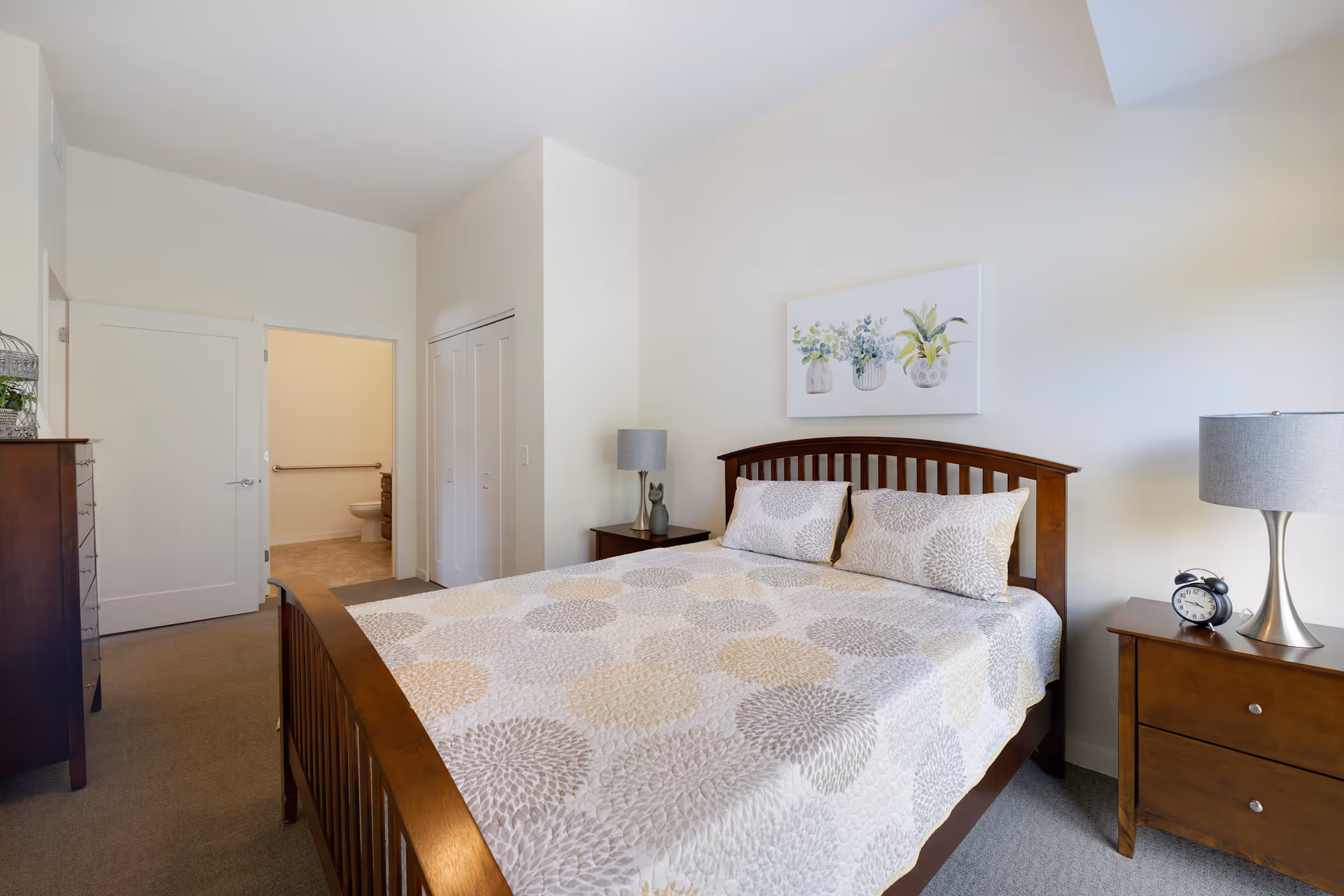 A bright bedroom with a wooden bed frame and a bed covered with a patterned quilt and two pillows. There are two wooden nightstands on either side of the bed, each with a lamp, and an alarm clock on the right nightstand. A painting of three potted plants hangs on the wall above the bed. A dresser is visible on the left side of the room. An open door reveals a bathroom with a toilet and grab bars.