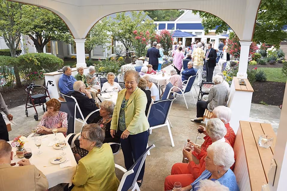 A group of elderly people socializing and enjoying drinks at an outdoor covered patio area with tables and chairs, surrounded by greenery and flowers at a retirement community.