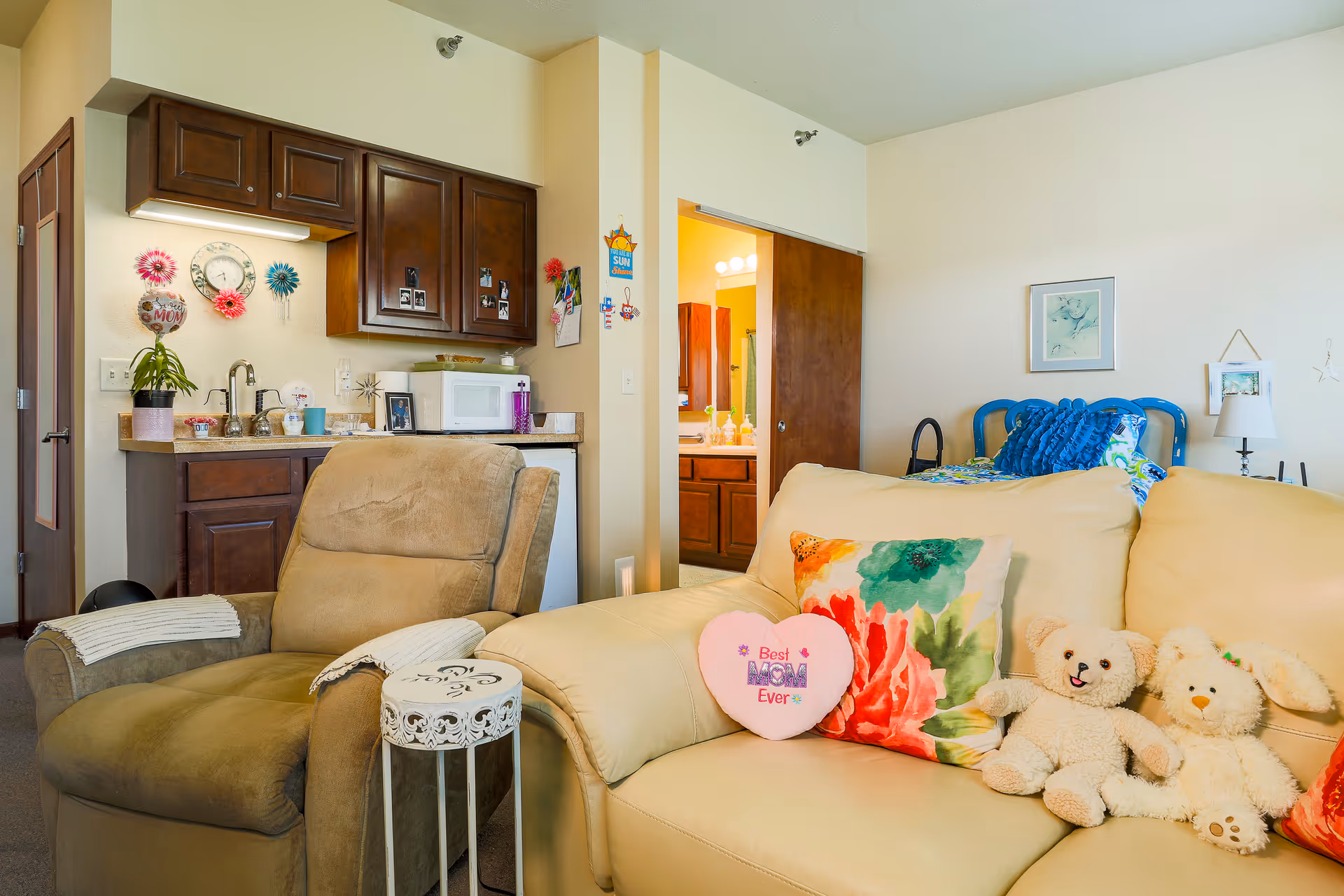 A cozy living space in a senior living facility featuring a beige leather sofa with colorful pillows and stuffed animals, a brown recliner chair with white arm covers, a small decorative side table, and a kitchenette with dark wooden cabinets, a microwave, and a sink. In the background, there is a bed with blue pillows and a framed picture on the wall, and an open door leading to a bathroom with a lit vanity mirror.