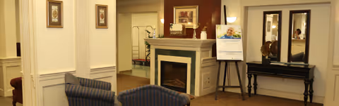 Cozy senior living lounge with armchairs, a fireplace, mirrors, and an information board.