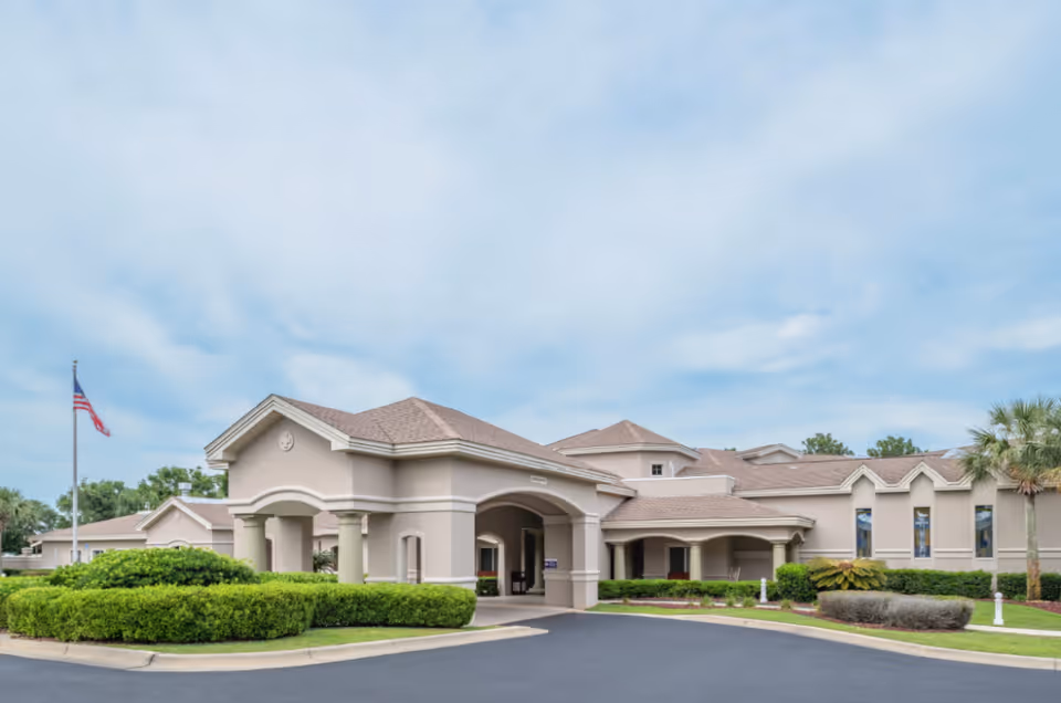 Exterior view of a single-story senior living facility building with beige walls and a brown roof. The entrance features a covered driveway with columns. There are well-maintained green bushes and palm trees around the building, and an American flag is visible on a flagpole to the left. The sky is partly cloudy.