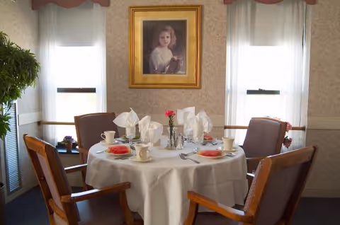 Round dining table set for four with a white tablecloth, napkins, cups and plates in a cozy room with a framed portrait between two windows.