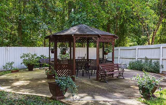 Outdoor seating area with a wooden gazebo surrounded by a white fence and greenery. The gazebo has a shingled roof and contains a table with chairs. There are benches and potted plants around the paved area.