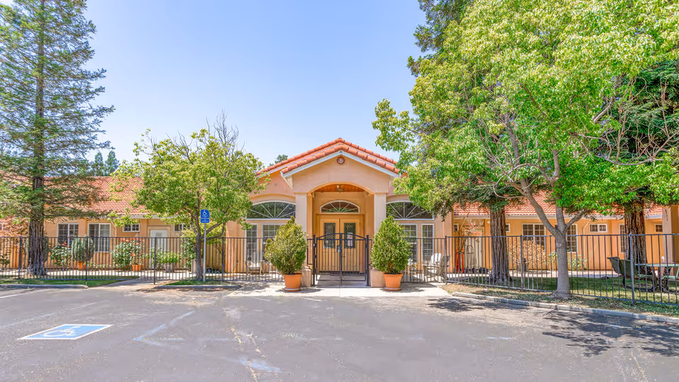 Front exterior view of Senior Care Villa of Loomis, showing a single-story building with a tiled roof, a gated entrance flanked by two potted plants, several trees, and a parking area with a handicapped parking space.