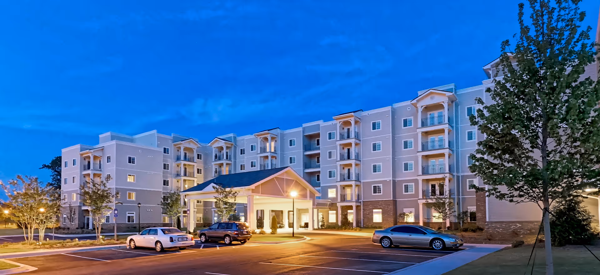 Exterior view of a multi-story senior living facility building at dusk with a covered entrance and several parked cars in the parking lot. The sky is clear with a deep blue color, and the building is illuminated with warm lights.