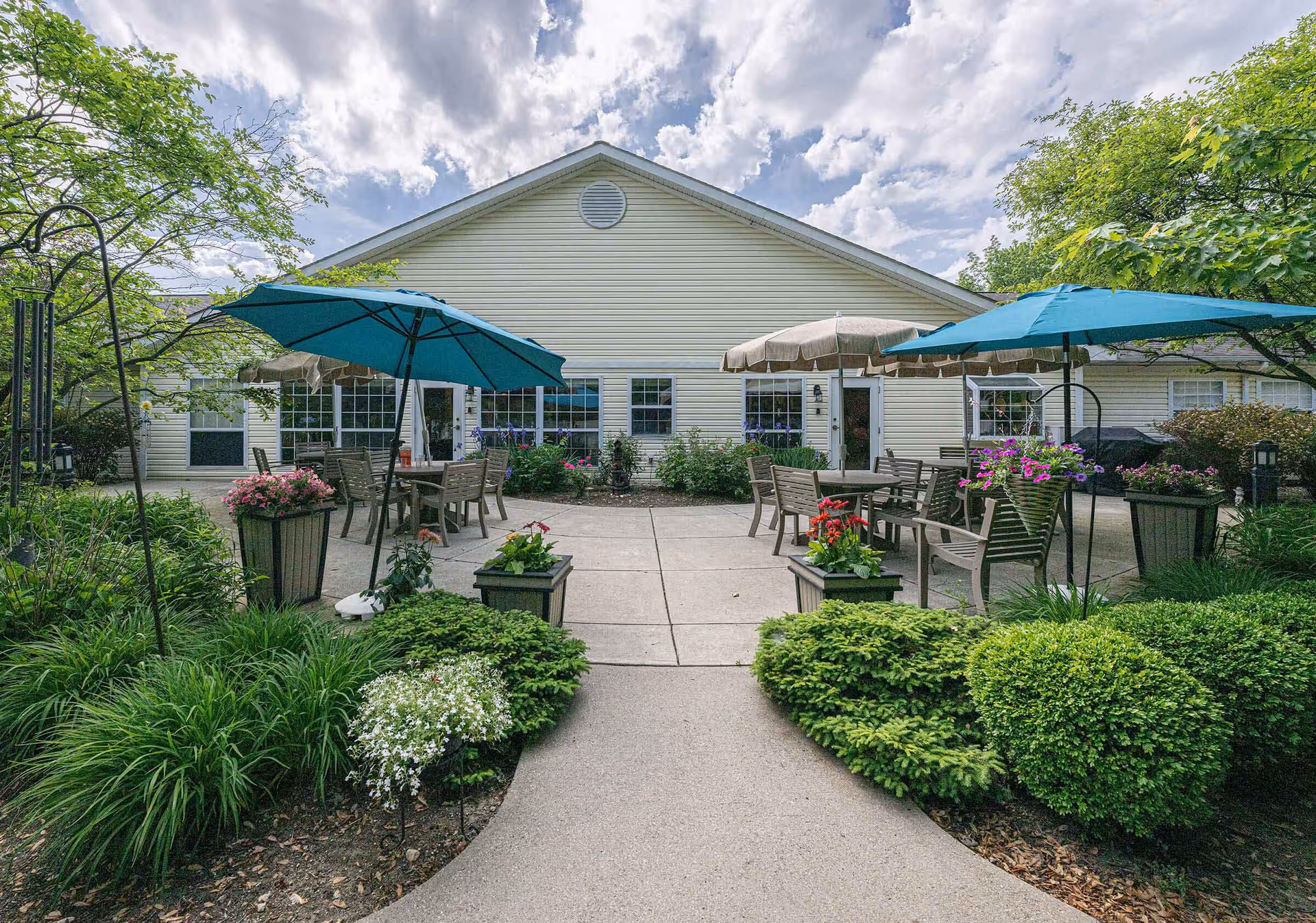 Outdoor patio area at Arden Courts - ProMedica Memory Care Community (Westlake) featuring several tables with chairs and large umbrellas surrounded by lush greenery and flowering plants, with a light-colored building in the background under a partly cloudy sky.