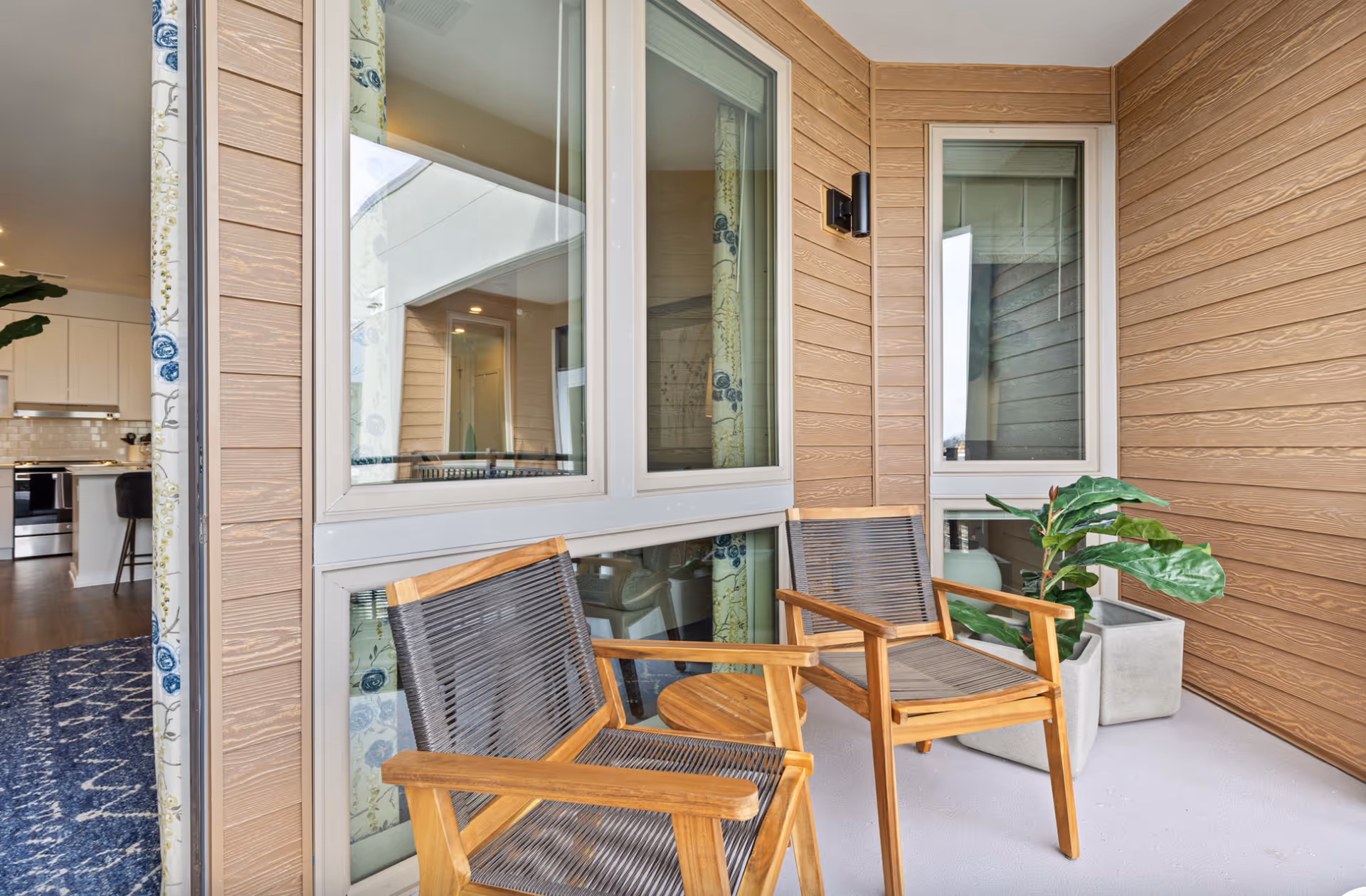 A small outdoor patio area with two wooden chairs featuring woven seats and backs, a small round wooden table between them, and two potted plants in square gray planters. The patio is enclosed by light brown wooden siding and large windows that look into an interior space with a kitchen and living area visible.