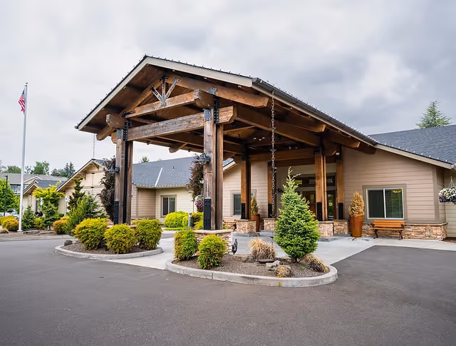 Exterior view of Sunnyside Meadows Memory Care facility showing a large wooden covered entrance with stone accents, surrounded by landscaped bushes and trees under a cloudy sky.
