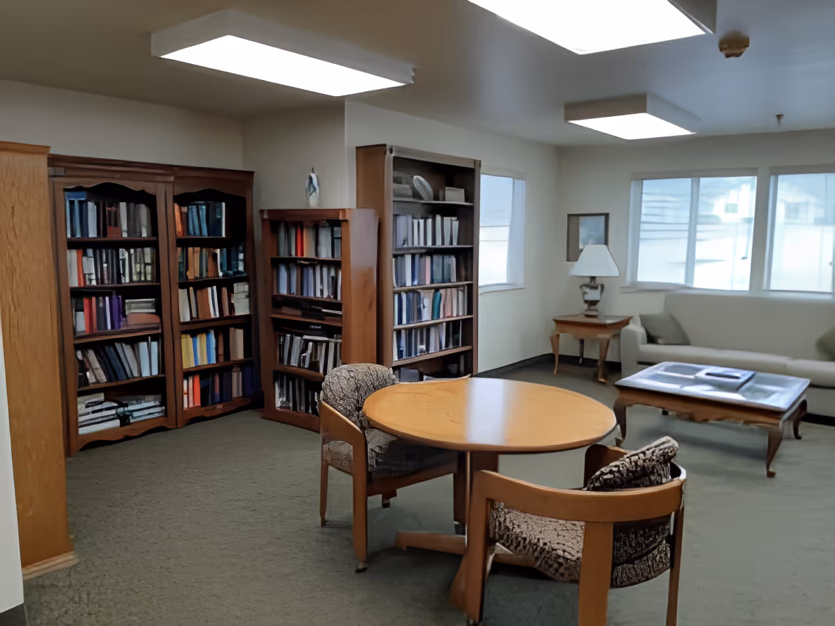 A quiet common room with bookshelves, a round table and chairs, a sofa, and a coffee table under ceiling lights.