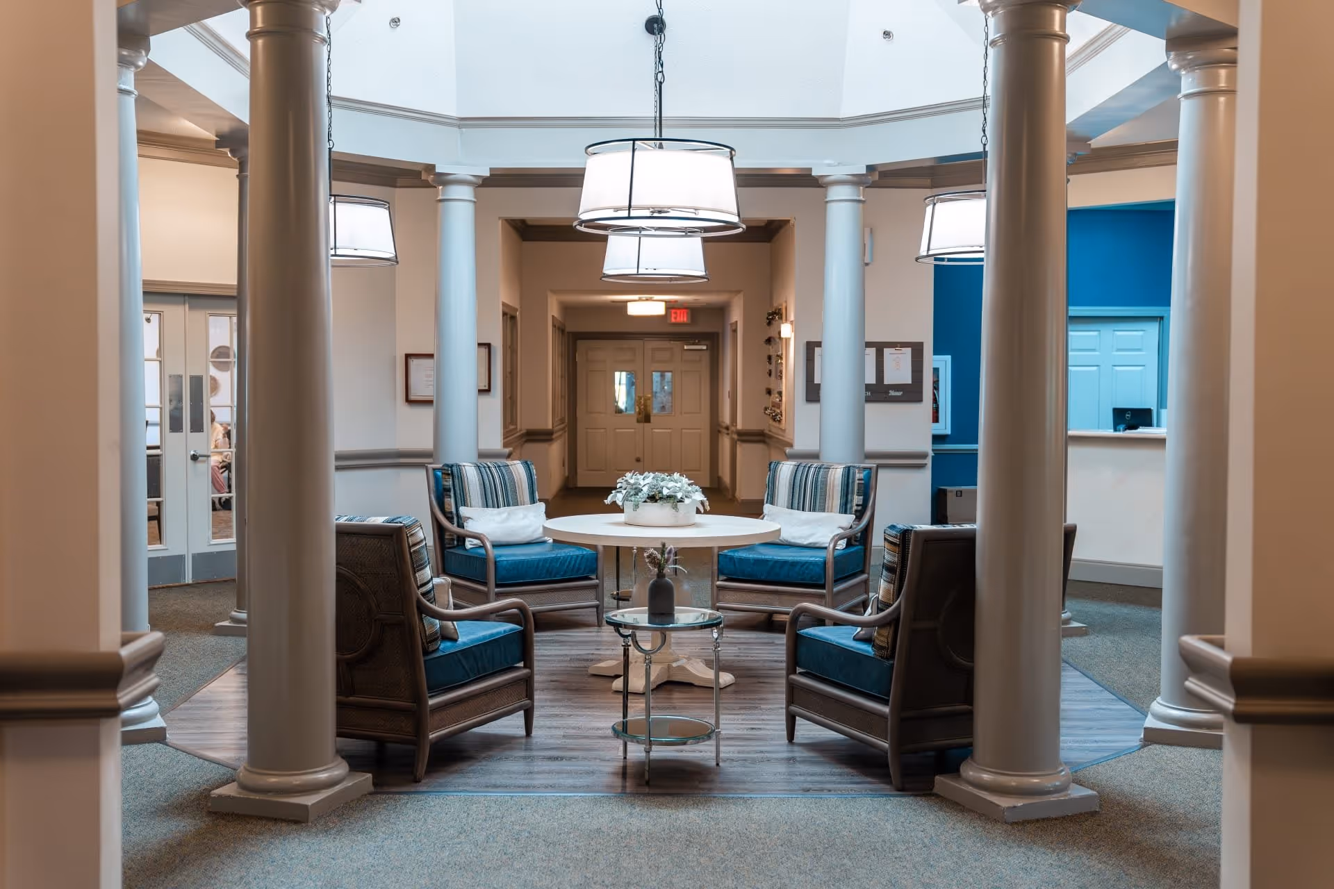 A cozy seating area in a senior living facility with four cushioned armchairs arranged around a round white table with a flower centerpiece. The space is surrounded by white columns and has soft lighting from hanging lamps. In the background, there is a reception desk with a blue accent wall and double doors leading to another room.