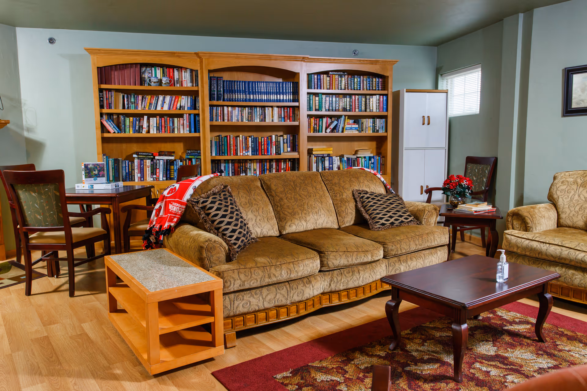Cozy living room in a senior facility with a patterned sofa and armchairs, a coffee table, bookshelves filled with books, and a small table and chairs.