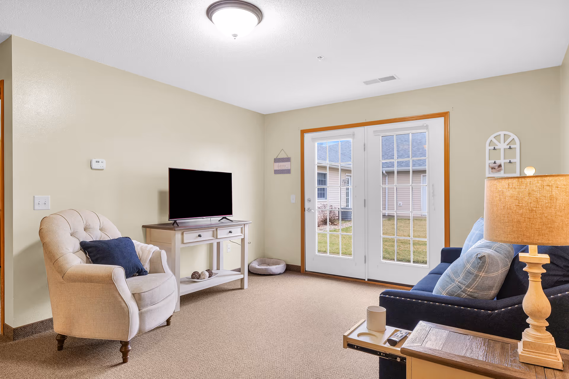 A cozy living room with beige walls and carpet. The room features a beige armchair with a dark blue pillow, a small wooden TV stand with a flat-screen TV, a dark blue couch with light blue pillows, a wooden side table with a lamp, and French doors leading outside to a grassy area.