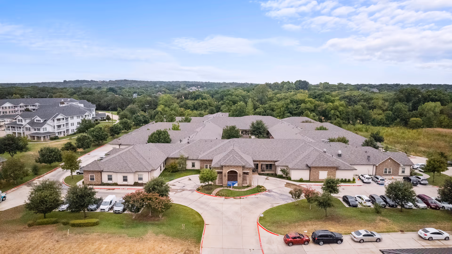 Aerial view of a single-story senior living building with a central entrance, surrounding parking, and landscaped grounds.